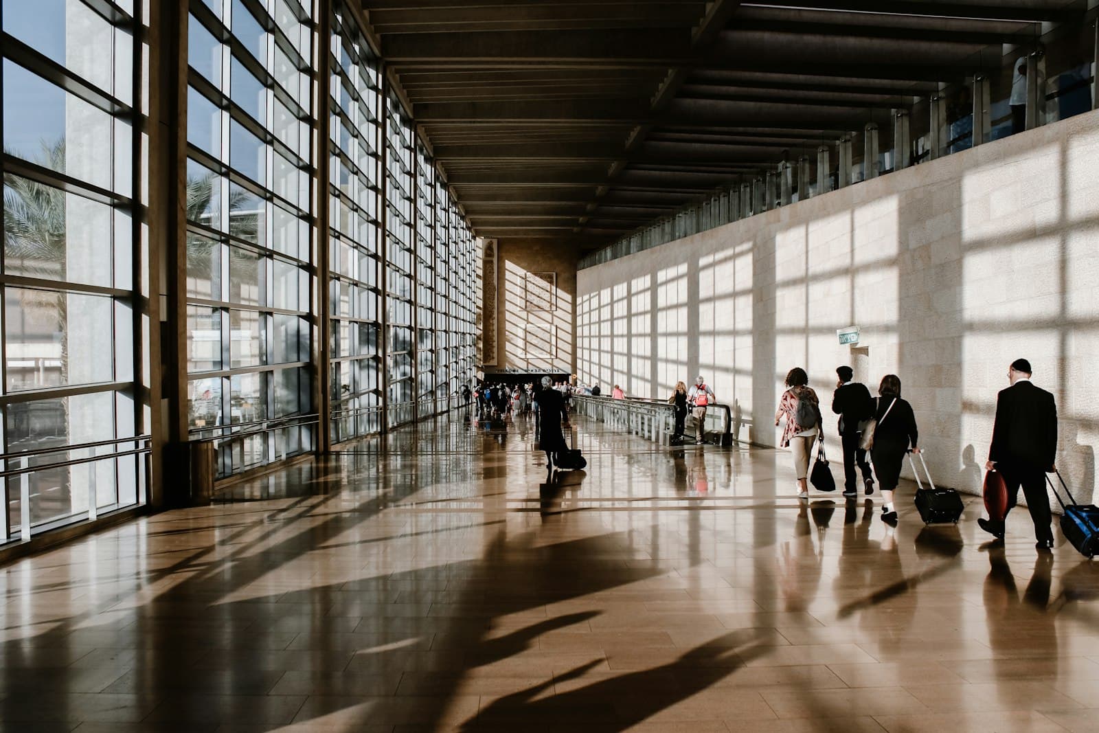 Travelers pulling luggage through a sunlit airport terminal corridor