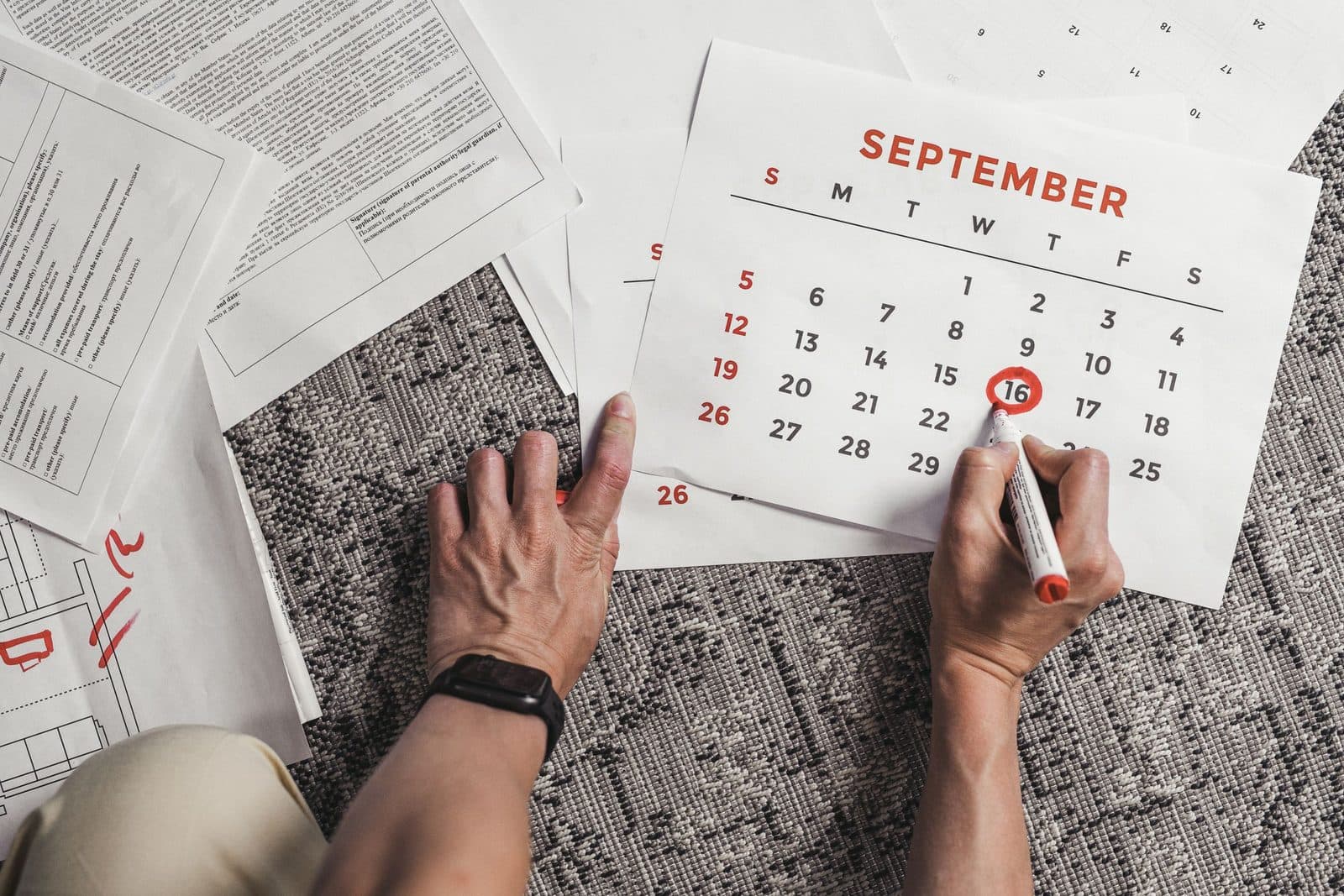 A hand marking a date on a September calendar surrounded by documents